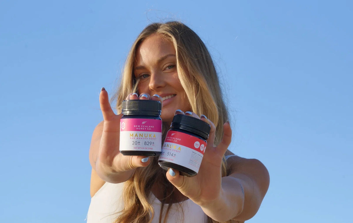 Woman holding two jars of Manuka honey products against a clear blue sky background.
