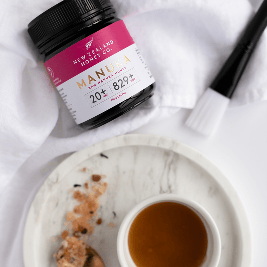 New Zealand manuka honey co. UMF 20+ manuka honey sitting bench top on crisp white cloth above a marbvle dish with manuka honey and other skincare ingredients with a face mask brush and
