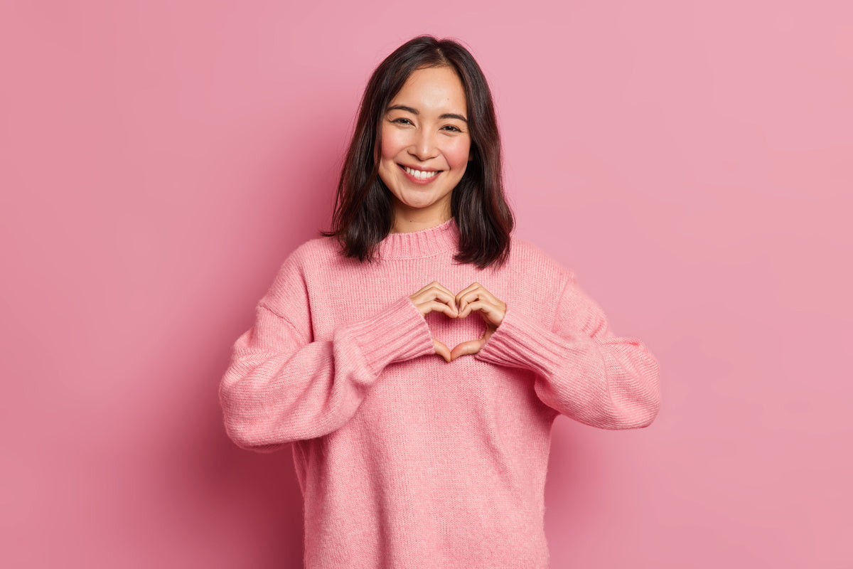Smiling person in a pink sweater making a heart shape with hands.