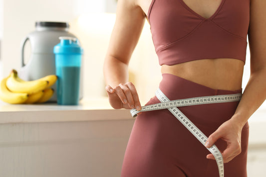 Person measuring waist with tape in fitness attire, bananas and water bottle nearby.