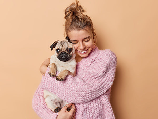 Woman hugging a pug dog against a light orange background.