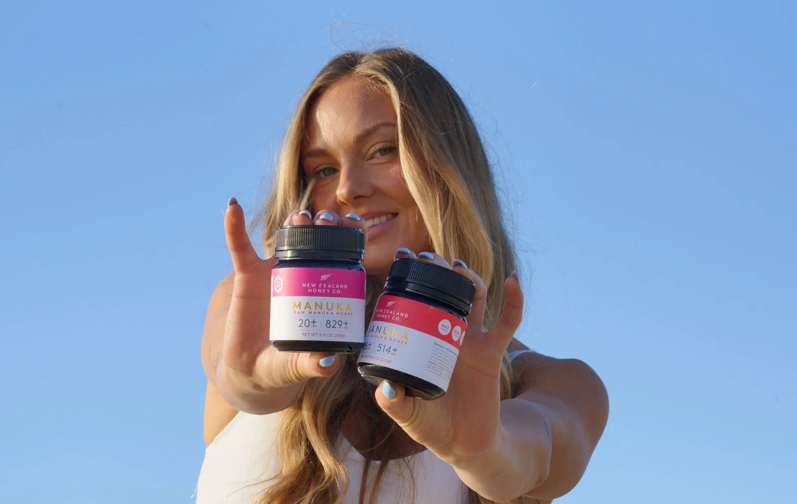 Woman holding two jars of Manuka honey products against a clear blue sky background.