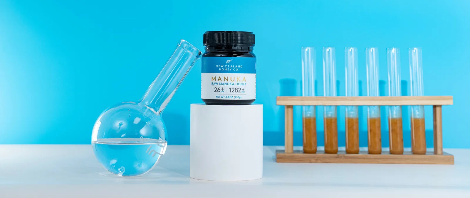 New Zealand Honey Co. manuka honey jar displayed with laboratory glassware and test tubes against a bright blue background.