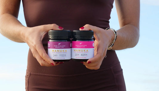 Person in brown workout attire holding two jars of New Zealand Manuka honey with pink labels showing different UMF ratings.