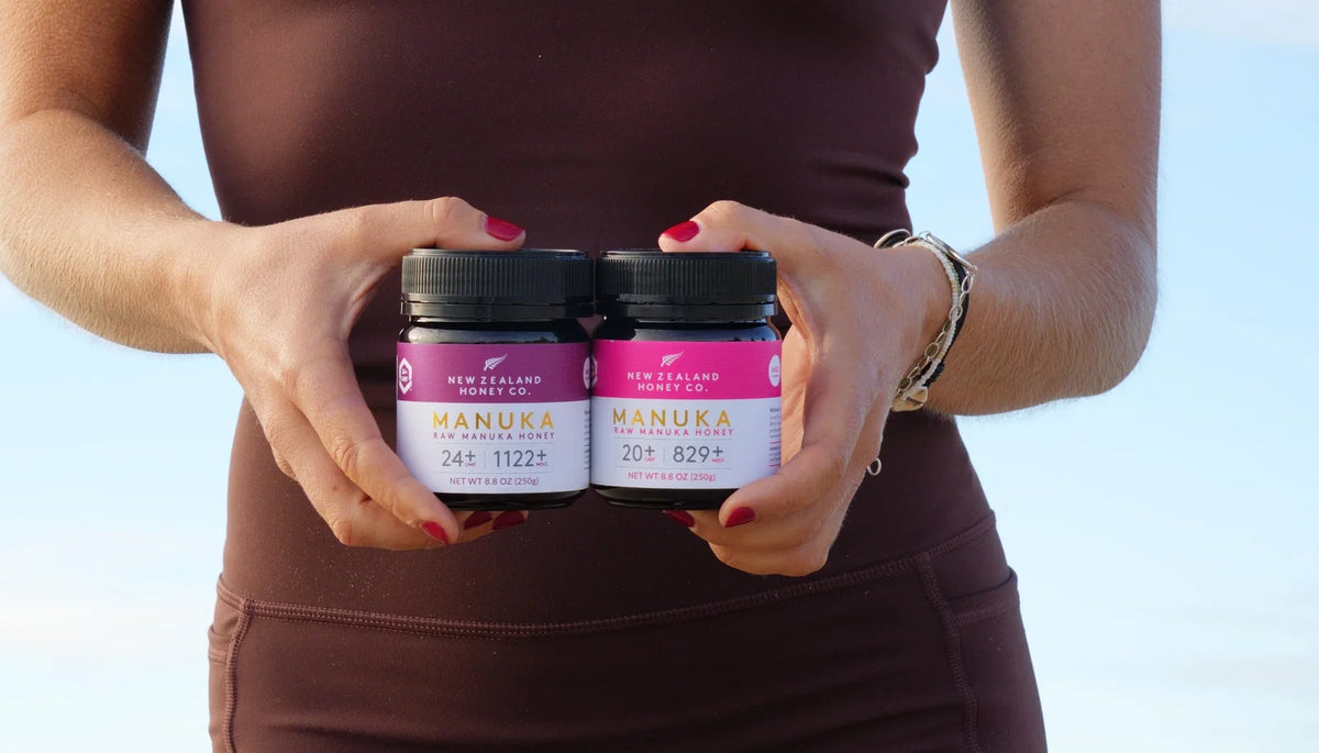 Person in brown workout attire holding two jars of New Zealand Manuka honey with pink labels showing different UMF ratings.