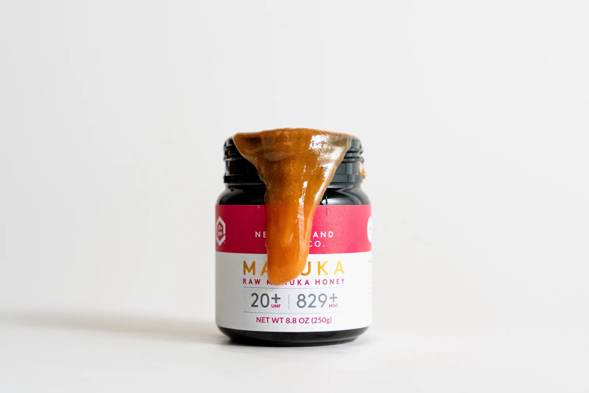 Jar of raw Manuka honey with UMF 20+ rating, honey dripping from spoon on top, pink and white label.
