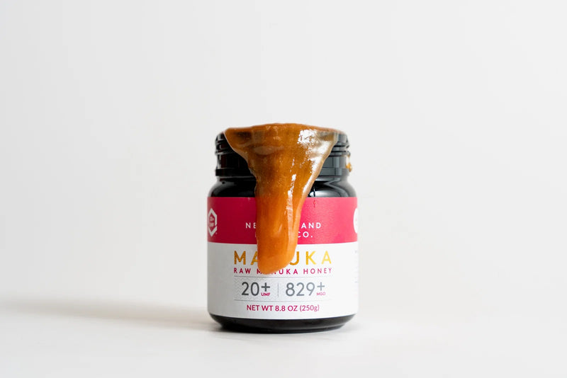 Jar of raw Manuka honey with UMF 20+ rating, honey dripping from spoon on top, pink and white label.