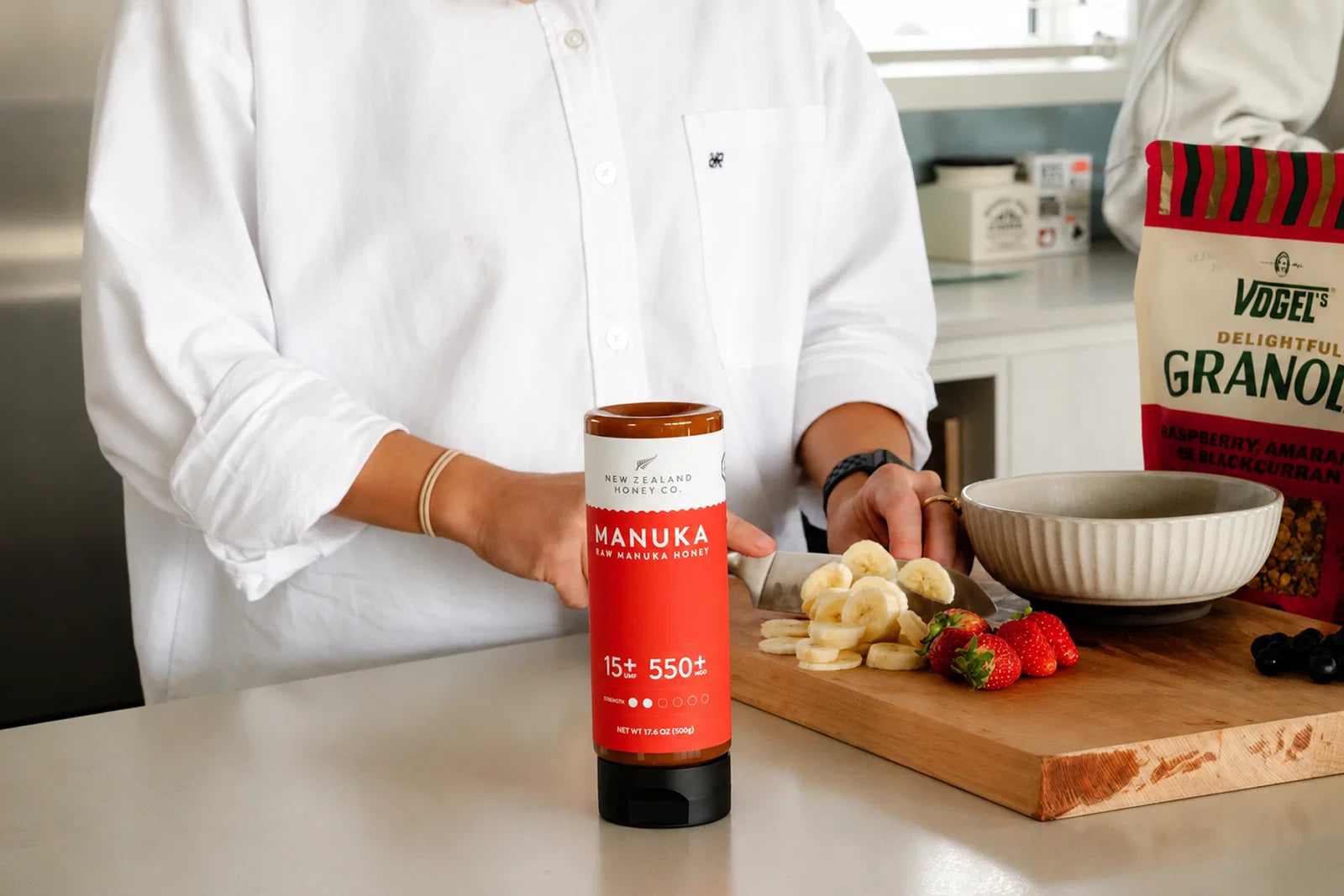 Person in white shirt holding Manuka honey jar beside wooden cutting board with fresh fruit and granola box