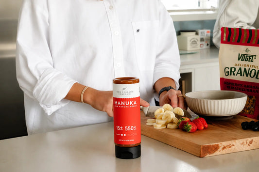 Person in white shirt holding Manuka honey jar beside wooden cutting board with fresh fruit and granola box