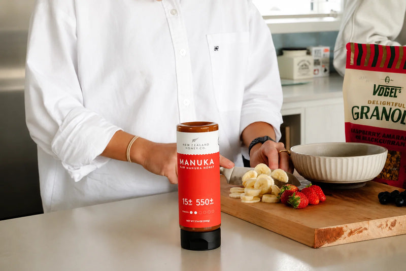 Person in white shirt holding Manuka honey jar beside wooden cutting board with fresh fruit and granola box