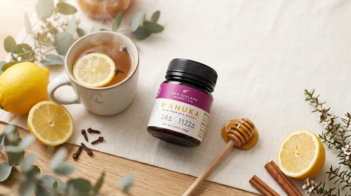 Manuka honey jar beside lemon tea, lemons, cloves, cinnamon, eucalyptus, and a honey dipper.