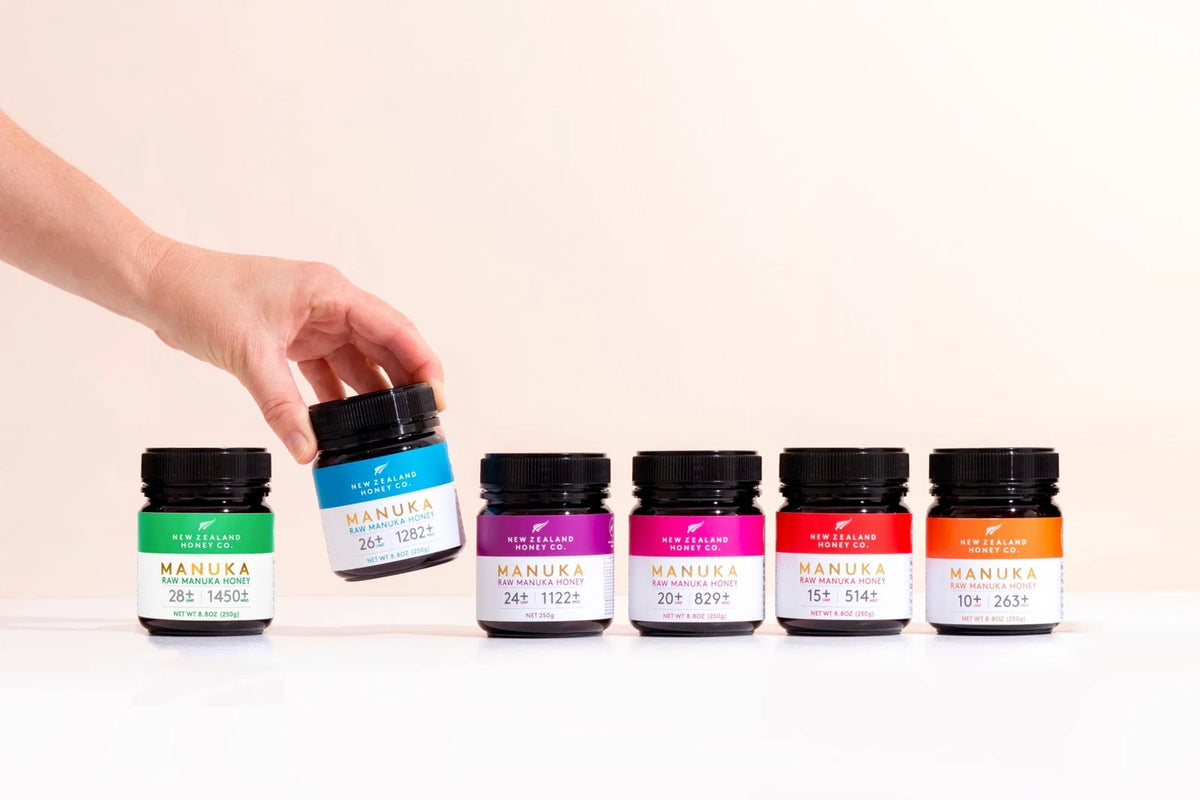 Hand holding a Manuka honey jar above five colorful Manuka honey jars arranged in a row.