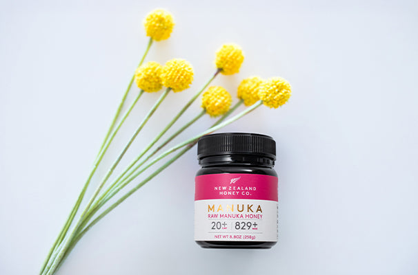 Jar of Manuka Honey beside yellow flowers on a light background.