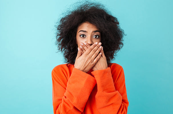 A surprised woman covers her mouth with her hands against a blue background.