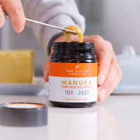 Hand using a spoon to scoop honey from an open New Zealand Honey Co. Manuka Honey UMF™ 10+ MGO 263+ jar on a kitchen counter