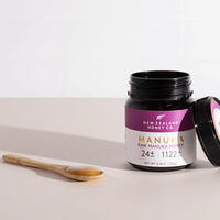 Open New Zealand Honey Co. Manuka Honey UMF™ 24+ MGO 1122+ jar tilted to show the lid, with a bamboo spoon beside it on a beige surface, 250g (8.8oz)