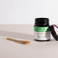 Open New Zealand Honey Co. Manuka Honey UMF™ 28+ MGO 1450+ jar with the lid leaning beside it, bamboo spoon to the left on a beige surface, 250g (8.8oz)