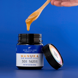 Open New Zealand Honey Co. Manuka Honey UMF™ 30+ MGO 1620+ jar with a wooden honey dipper drizzling honey from above on a deep blue background