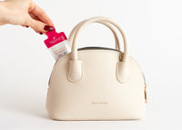 Hand placing a single New Zealand Honey Co. Manuka Honey on-the-go packet UMF™ 20+ MGO 829+ into a small beige handbag — lifestyle scene