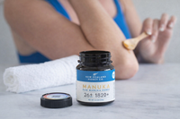New Zealand Honey Co. Manuka Honey Mini jar (blue UMF 26+ label) on a bathroom counter beside a rolled towel and lip balm — face-mask lifestyle scene